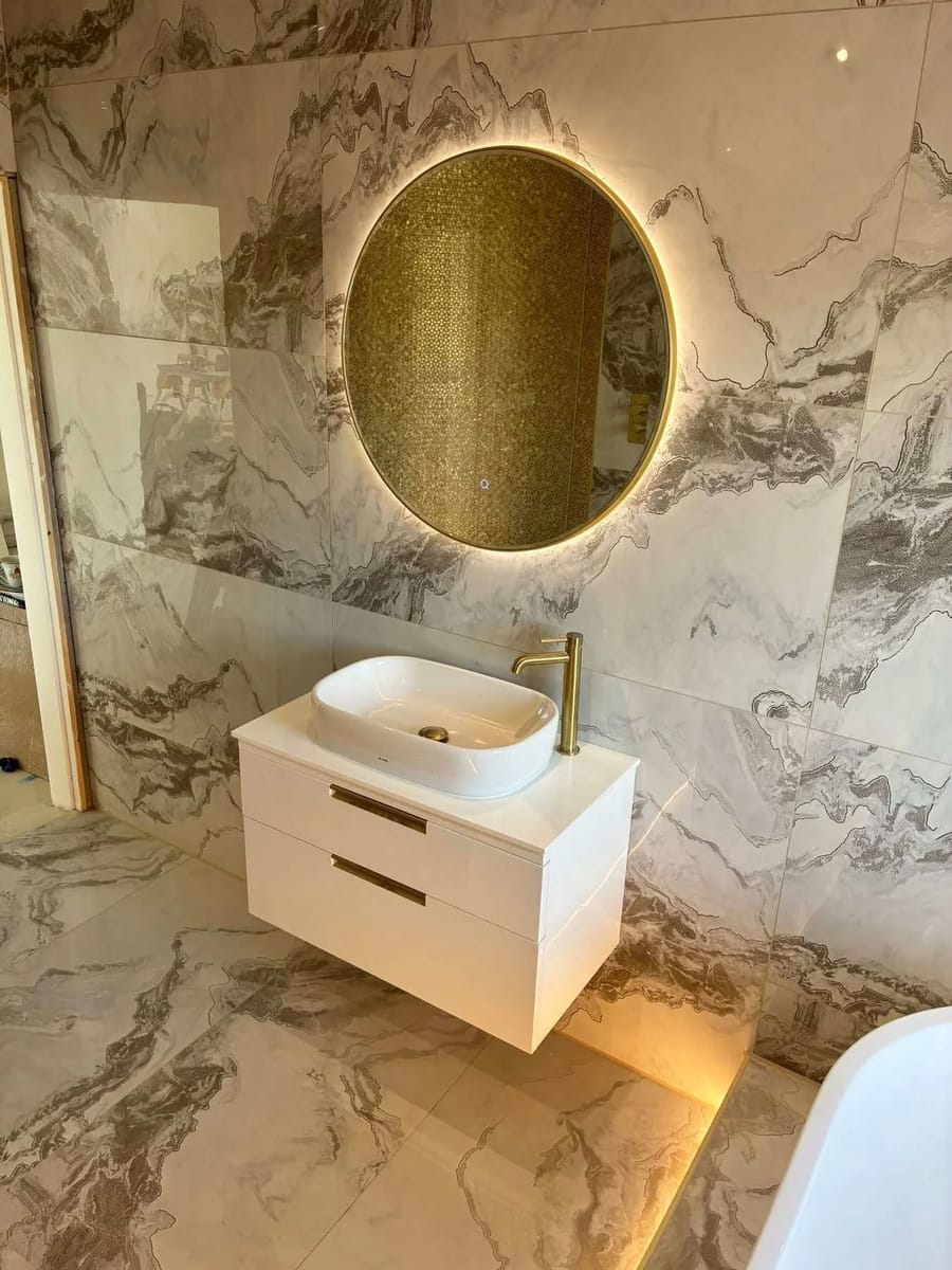 A marble tiled bathroom with a gold tap and a backlit round mirror, fitted by Askern Plumbing